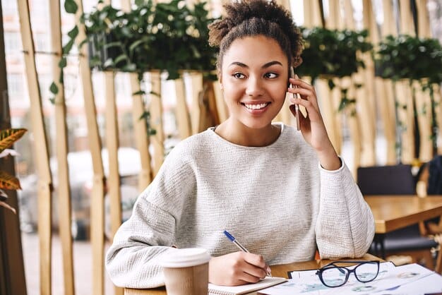 A person confidently talking on the phone, smiling and taking notes.