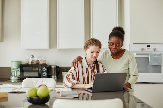 A parent and a teenager sitting together at a kitchen table, looking at a laptop. The laptop screen displays a budgeting app. Both are smiling and pointing at the screen, indicating a collaborative and positive learning experience. The kitchen is modern and well-lit.